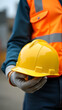 © narumon - Close-up of construction worker hand holding yellow safety helmet, leather gloves and orange vest. Workplace safety concept, vertical shot.