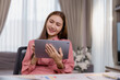 © Tj - Young asian businesswoman working from home, sitting at a desk and using tablet, smiling and looking at the screen, with documents and charts on the desk