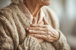 © QQPK14 - Elderly person clutching their chest, suggesting discomfort or pain, wearing a knitted sweater with a soft, neutral background emphasizing concern and vulnerability