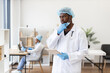 © sofiko14 - Male adult healthcare professional in medical office wearing scrubs, blond hairnet, gloves and stethoscope, holding clipboard, contemplating work with background colleague on laptop.