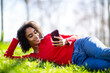 © mimagephotos - Young Black woman lying sideways on grass with smartphone, relaxed and smiling.