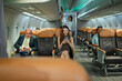 © Wanwajee - Wide side view of airplane cabin with female passenger using smartphone, other travelers seated nearby, and flight attendant checking overhead bins, concept of aviation safety and modern air travel.