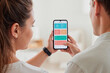 © DragonImages - Young adult Caucasian woman and young adult Caucasian man using smartphone together, woman holding phone displaying health tracking app with charts and statistics visible on screen