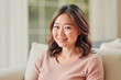 © DragonImages - Portrait of young adult Asian woman smiling while sitting on sofa, looking into camera with relaxed expression, medium length hair styled in loose waves, indoor setting with soft background