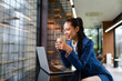 © amnaj - Asian businesswoman drinking coffee while working on her laptop in a modern office, enjoying a break and smiling