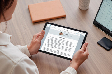 Wall Mural - Caucasian young adult woman holding digital tablet reading online document at desk, hands visible gripping device, laptop and notebook on wooden table, business setting