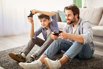 Videogames concept. Joyful father and son competing in online games, holding joysticks while sitting on carpet in living room and spending time together on weekend, copy space