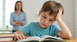 © Cleverson - Frustrated boy doing homework with a mother watching over him in the background. He looks stressed and overwhelmed while studying.