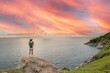 © Sirirat Makprasert - Woman Standing on Rock Cliff Watching Sunset Over Sea, A lone traveler standing on a cliff rock, gazing at the glowing red-orange sunset sky over the wide ocean, symbolizing adventure.