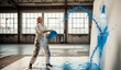 © Paylopi - An artist in a messy, paint-splattered studio throws a bucket of blue paint at a large white canvas, a dynamic and expressive act of creating abstract art.