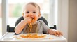 © Aminul - Cute baby enjoying spaghetti lunch sitting in a high chair with a messy face and hands eating noodles with tomato sauce in a bright indoor setting