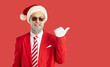 © Studio Romantic - Cheerful, funny, smiling, happy, young Santa Claus in sunglasses, dressed in red suit and warm Santa hat, points with his thumb at the copy space. Studio portrait on isolated red background.