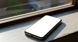 © Djemari - A modern smartphone rests on a windowsill, bathed in sunlight, showcasing a blank screen.
