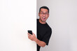 © KrishnaTedjo - Adult Asian man holding mobile phone peeking from behind wall with wow face expression