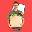 © New Africa - Delivery service worker giving order out of mobile phone against red background. Man with paper bag full of food products