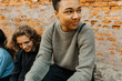© Maskot - Young man looking away while sitting with friends in front of brick wall