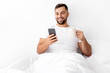 © Prostock-studio - Cheerful young muscular caucasian male drinking coffee chatting on smartphone and enjoy good morning, weekend in bedroom interior. New app, advertising and offer, communication remotely, empty space
