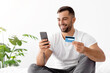 © Prostock-studio - Smiling european handsome young guy in white t-shirt shopping online, orders goods on smartphone and shows credit card in bedroom interior. Payment money and buy purchase, sale and shopaholic at home