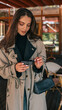 © shine.graphics - Stylish young woman in beige trench coat smiling while holding a small matte black gift box with satin ribbon, standing outdoors near cafe tables