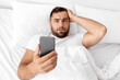 © Prostock-studio - Despaired upset millennial european muscular man lies on white bed holds head with hand and reads news on smartphone in bedroom, top view. Facial expression difficulties, stress, problems and bad news