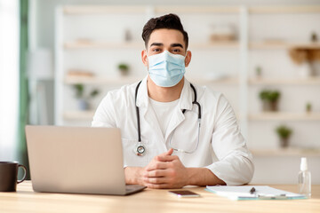 Doctor in protective face mask sitting at desk and working on laptop at clinic, having appointments during coronavirus pandemic. Male doc sitting at cabinet in hospital, copy space