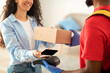 © Prostock-studio - Happy woman using smartphone for contactless payment, black male courier wearing protective gloves and thermo backpack holding Point Of Sale machine and cardboard box with purchase