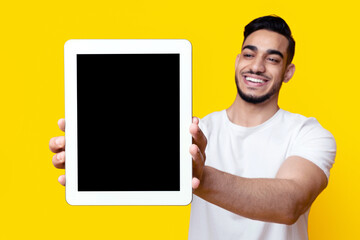 Cheerful handsome middle-eastern young man showing modern digital tablet with empty screen, recommending nice mobile application or showing amazing offer over yellow studio background, mockup