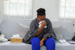 © David - African American woman wrapped on a couch,in a blanket, with flu virus blowing her nose at home.She appears to be sick or to have allergies.