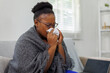 © David - African American woman wrapped on a couch,in a blanket, with flu virus blowing her nose at home.She appears to be sick or to have allergies.