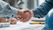 © CStock - Two individuals shaking hands over a table covered in blueprints and construction materials, symbolizing collaboration.