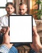 © Dzzle - View of hands holding a blank tablet, with two blurred men on a couch in the background, lit by warm indoor light