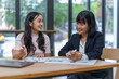 © amnaj - Two Asian businesswomen analyzing financial charts and engaging in discussion during a corporate meeting, fostering teamwork and collaboration