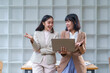 © amnaj - Two Asian businesswomen engaging in a work discussion while using a laptop in a modern office environment, showcasing collaboration and teamwork