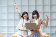 © amnaj - Two happy asian businesswomen raising arms and celebrating success while looking at laptop in the office
