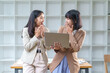 © amnaj - Two Asian businesswomen standing at a desk in a bright office, looking surprised while analyzing data on a laptop during a meeting