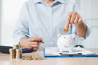 © doidam10 - Concept of saving money and investment planning. A businesswoman drops coin into white piggy bank on office desk with documents, symbolizing financial security, budget management and future wealth.