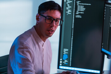 Programmer wearing glasses codes at dual monitors in modern office, lit by cool blue light. Scene highlights developer focus, software engineering, technology, and computer workstation setup.