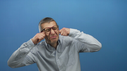 Man wearing glasses mimics crying in front of a blue wall using his fists near eyes while dressed in a striped shirt.