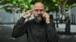 © Krakenimages.com - Man with beard talking on phone outdoors in urban setting with serious expression, tattoo visible, surrounded by blurred trees in the background.
