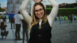 Woman smiling with heart gesture at pisa tower in italy, wearing glasses and sweater outdoors on a sunny day, surrounded by tourists enjoying the iconic landmark.