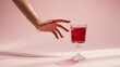© Jubida - Elegant womans hand reaching for a glass of red wine on a pink background in a studio setting