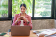 © bunyiam - A woman is sitting at a desk with a laptop and a cup of coffee