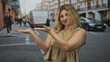 Woman blonde palms up shrug on busy city street showing open hands and casual smile while facing camera; curiosity doubt.
