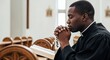 © David Zarzosa - Black priest praying with rosary in church pew beside open Bible with copy space.