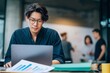 © id512 - Young asian man working on laptop with financial charts on desk in modern office. Casual employee or startup analyst focusing on business project while colleagues brainstorm in blurred background.