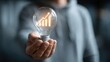 © Oulaphone - A person holds a glowing light bulb with an upward graph inside, symbolizing innovation and growth.