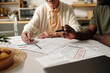 © pressmaster - Senior Caucasian woman and Black man reviewing financial documents together at table, discussing bills and payments, hands pointing at paperwork with overdue notice visible