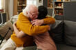 © pressmaster - Senior Caucasian man embracing young adult woman on sofa, both sitting closely and hugging warmly, walking cane visible beside man, home interior background with shelves and plants