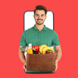© New Africa - Delivery service worker giving order out of mobile phone against red background. Man with crate full of food products