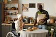© pressmaster - Senior Caucasian woman with disability sitting in wheelchair at table smiling while Black man bringing groceries and bread, both interacting in modern kitchen setting